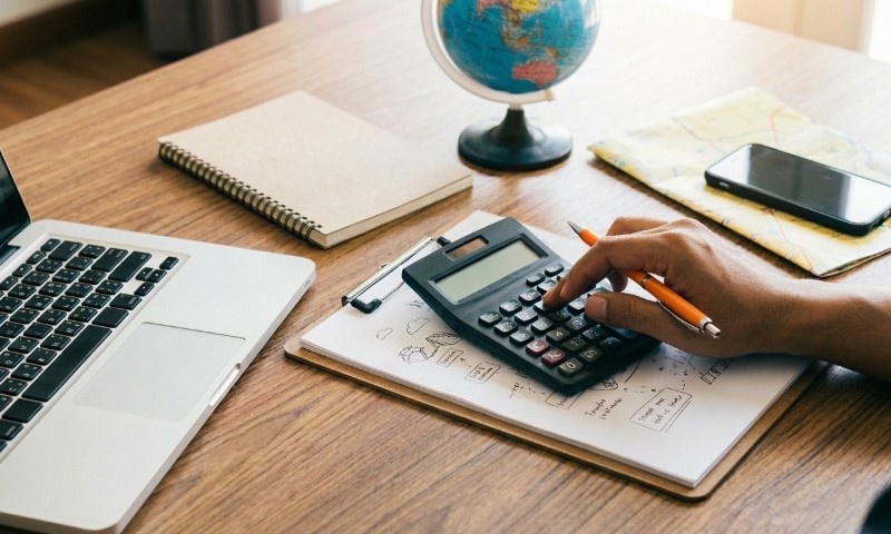 A hand uses a calculator on a wooden desk with a laptop, notebook, globe, and smartphone