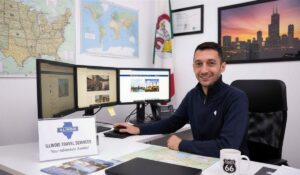 Travel Agent in Illinois sitting at a desk with three computer monitors, maps on the wall, and an Illinois Travel Services sign in the office